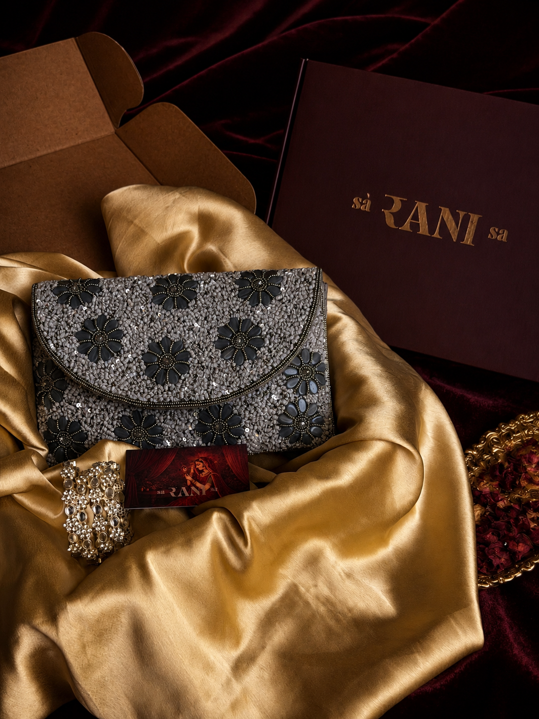 A close-up styled product shot of a dark floral clutch or envelope-style bag resting on draped gold satin fabric, with part of a brown box, a dark maroon branded box reading 'Saranisa', and a jeweled accessory visible around it.