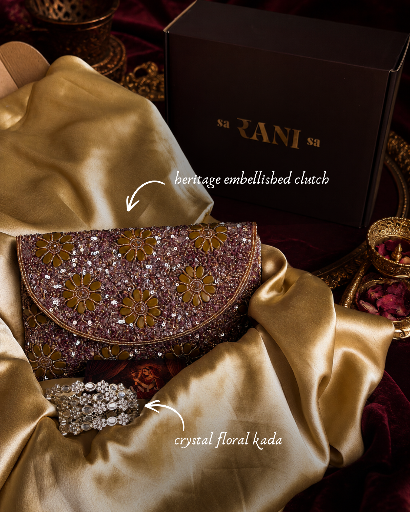 A close-up styled product shot of a brocade embellished clutch purse with floral embroidery, placed on draped champagne satin fabric beside a dark branded box and a crystal floral kada. Text labels identify the clutch and kada.