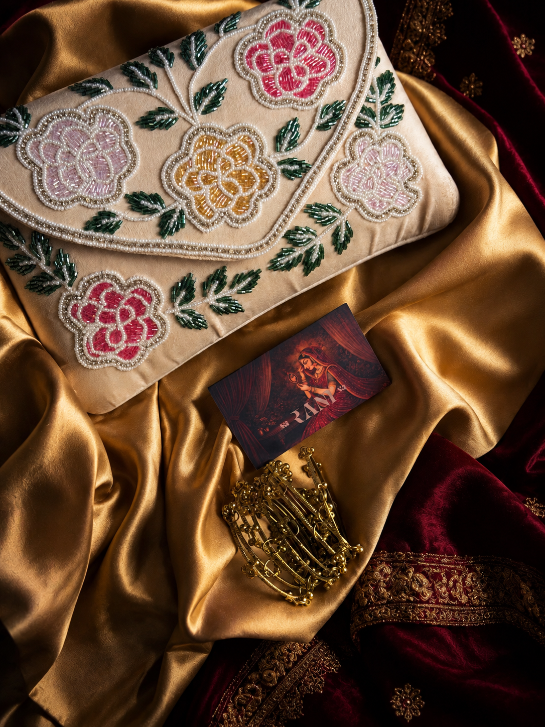An embroidered clutch bag with floral patterns in pink, yellow, and green on a beige base, styled on draped gold satin fabric alongside a gold bracelet and a small printed card.